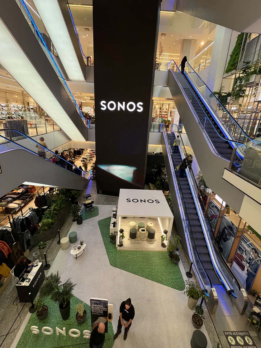 Vista en altura del stand Sonos montado en la tienda: alfombra verde, la isla de parlantes bajo el letrero SONOS y el tótem del concurso junto a las escaleras mecánicas