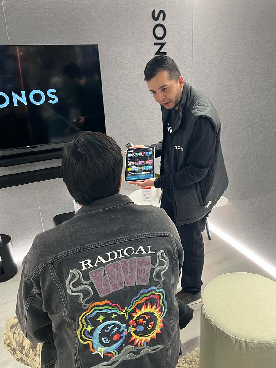 Un anfitrión del stand muestra en el iPad la pantalla de selección musical a un participante sentado frente al televisor