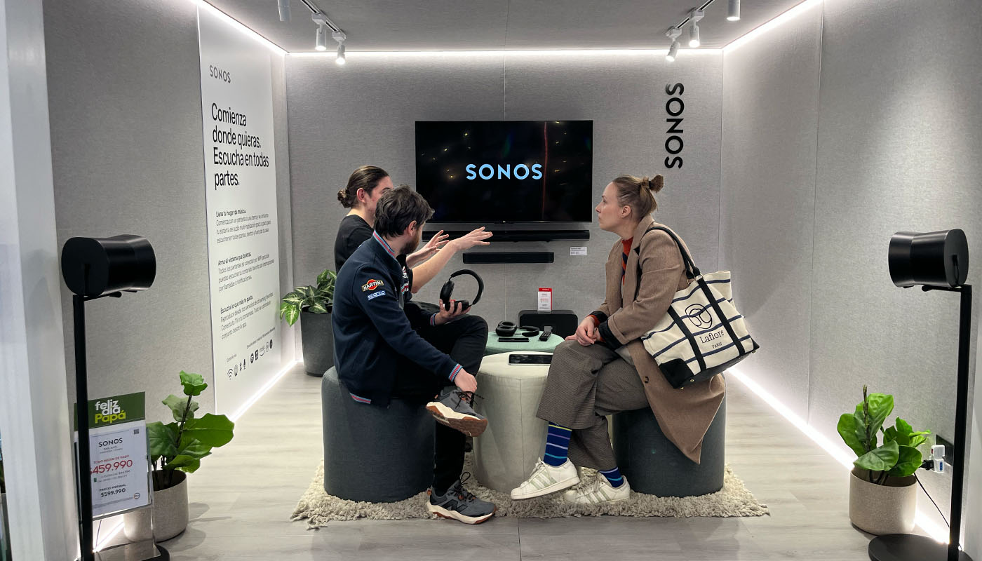 Un anfitrión del stand conversa con dos visitantes sentados frente al televisor en la sala de escucha, rodeados de parlantes y audífonos Sonos
