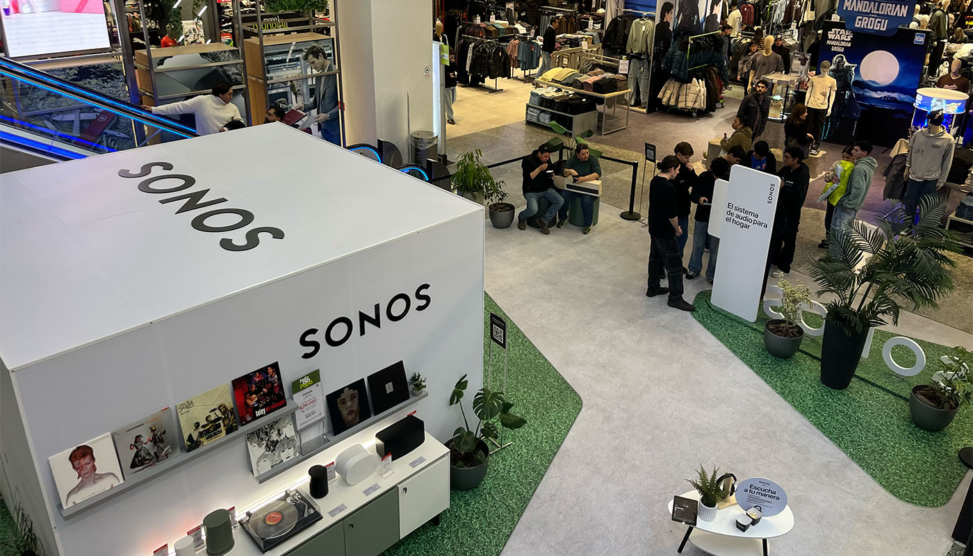 Vista en altura de la sala de escucha cerrada dentro de la tienda: la vitrina de vinilos y parlantes por fuera, y público esperando su turno junto al tótem del concurso